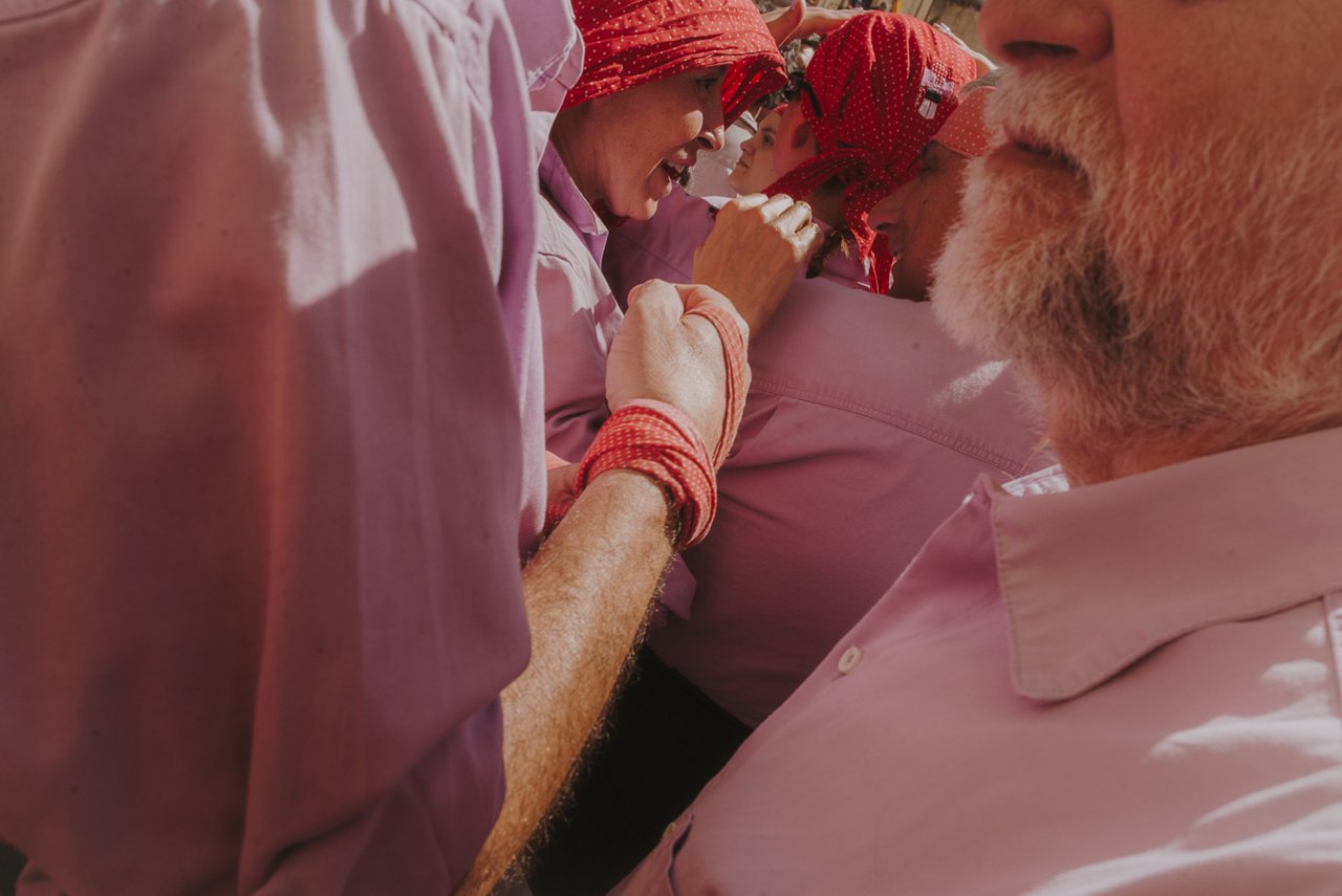 Close-up of several castellers wearing pink shirts and red headscarves and wristbands, standing very close together and holding on tightly to form the base. Their faces show concentration and effort, conveying cohesion, coordination, and intensity during the performance.
