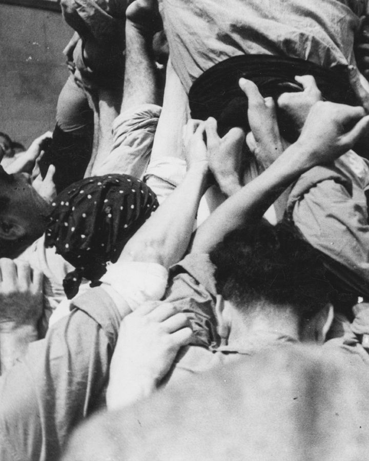 In black and white, a group of people standing very close together lift a casteller from the base. Several hands intertwine and support the body firmly, conveying collective effort and concentration in a historical scene of popular tradition.