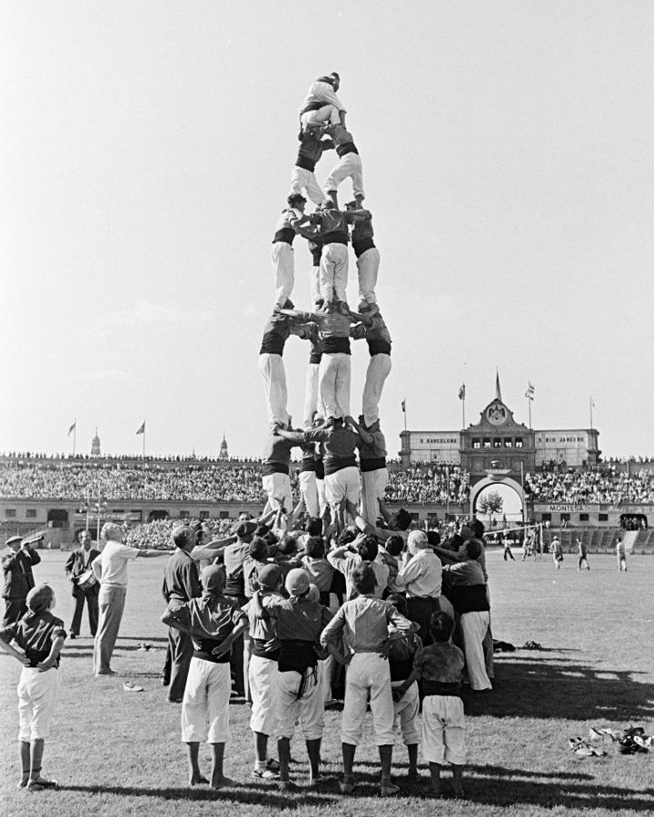 In black and white, a multi-level castell raised in an open space, surrounded by an audience. At the base, a large group of people supports the structure while the castellers climb to the top. In the background, a large grandstand filled with spectators and a building with a central arch can be seen, in a scene that conveys tradition, balance, and collective strength.