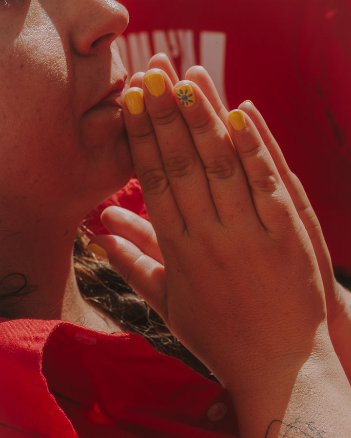 Close-up of a woman wearing a red shirt with her hands together near her mouth, as a sign of concern or anticipation. She has yellow-painted nails, with a floral detail on one of them, and a small tattoo on her wrist. The scene conveys concentration and support during a collective performance.