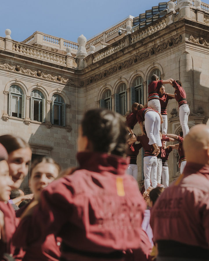 Castellers wearing maroon shirts and white trousers form a human tower in Plaça de Sant Jaume, in front of a historic building with an ornate façade and shuttered windows. Several levels of the structure are visible as the audience watches from below, in a scene that conveys tradition, balance, and teamwork.