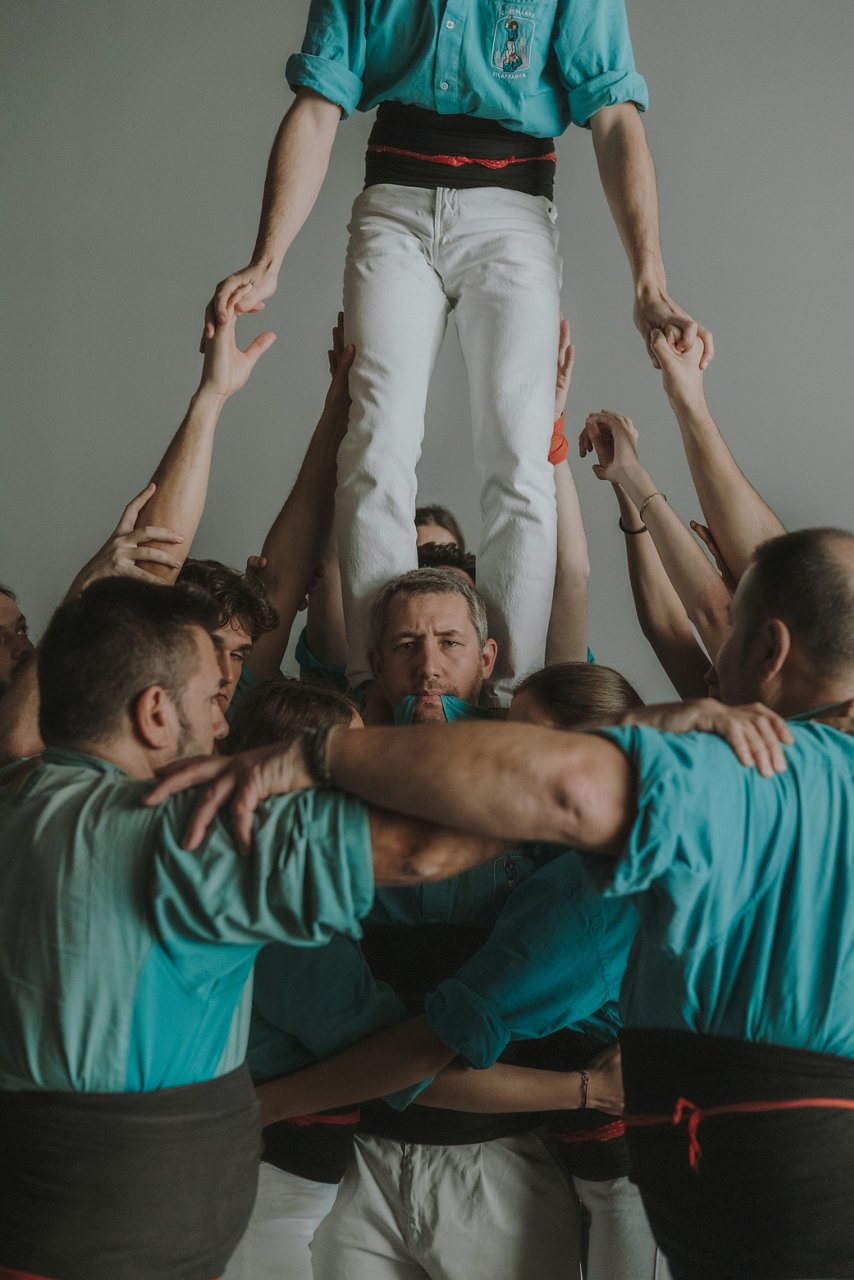 A group of castellers wearing green shirts form the base of a human castell while supporting a teammate climbing above them. Several hands help him maintain balance, and at the center a man with a focused expression can be seen holding the shirt lapels in his mouth. The scene conveys strength, coordination, and teamwork in a neutral setting.