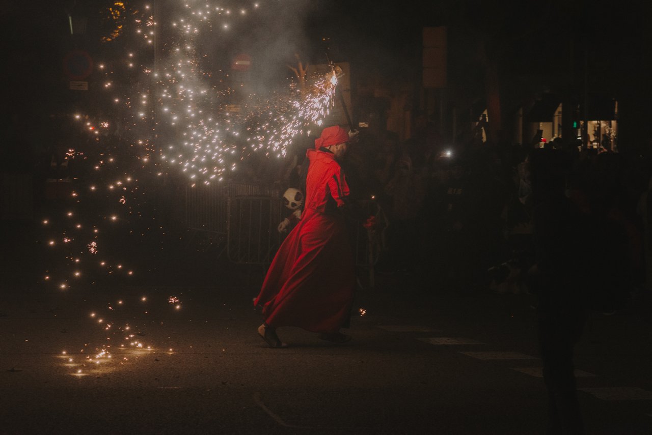 Une figure vêtue de rouge participe à un correfoc nocturne, courant dans la rue tout en tenant un élément pyrotechnique qui projette des étincelles autour d’elle. La scène est sombre et remplie de fumée et de lumière, avec le public en arrière-plan observant, et transmet énergie, intensité et tradition festive.