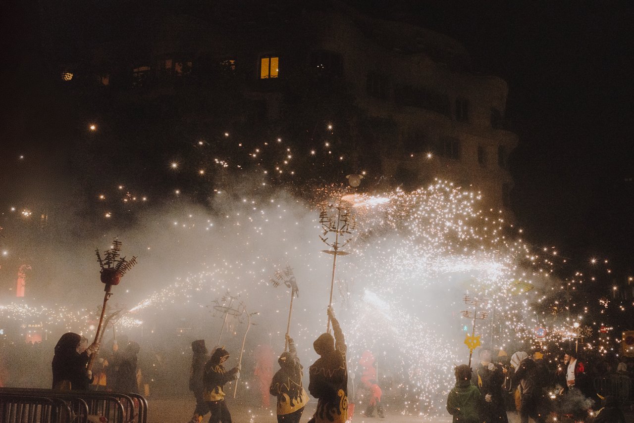 Un groupe de personnes participe à un correfoc nocturne en tenant des structures de feux d’artifice projetant des étincelles dans toutes les directions. La scène est remplie de fumée et de lumière, avec des bâtiments sombres en arrière-plan, transmettant intensité et tradition festive.