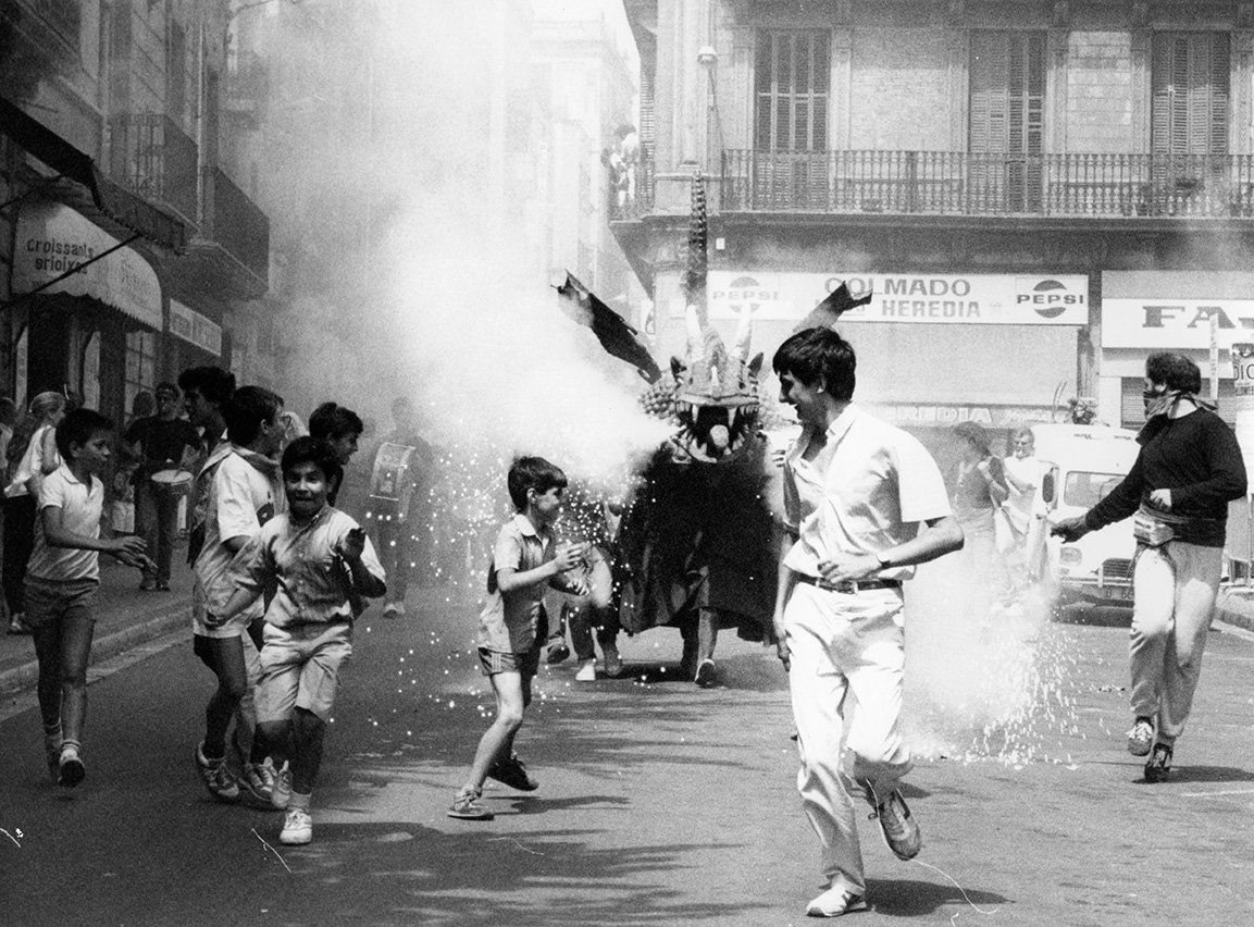 En noir et blanc, un correfoc sur une place de la Vila de Gràcia, avec des enfants et des adultes courant parmi les étincelles et la fumée tandis qu’une figure de diable avec des feux d’artifice avance au centre. La scène transmet mouvement, énergie et tradition populaire.