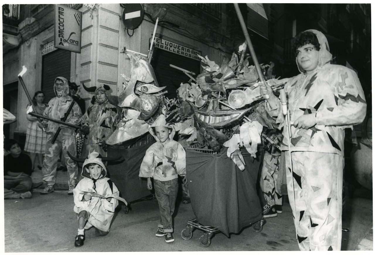 In black and white, a group of people, including children, dressed in devil costumes during a street festival. At the center, there are festive figures and structures shaped like heads and decorative elements, while participants hold torches and fire elements.