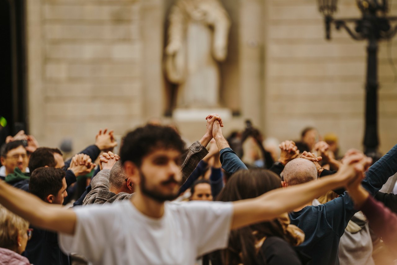 Grup de persones en una plaça amb els braços alçats i entrellaçats, ballant sardanes. En primer pla, un jove amb samarreta clara estén els braços, mentre al fons es veu una multitud concentrada.