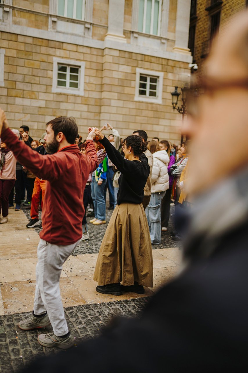 Persones en una plaça ballant sardanes amb els braços alçats i entrellaçats. En primer terme, un home amb camisa vermella i una dona amb faldilla llarga, mentre al fons un grup de gent forma una rotllana davant d’un edifici. Una figura desenfocada en primer pla aporta profunditat. 