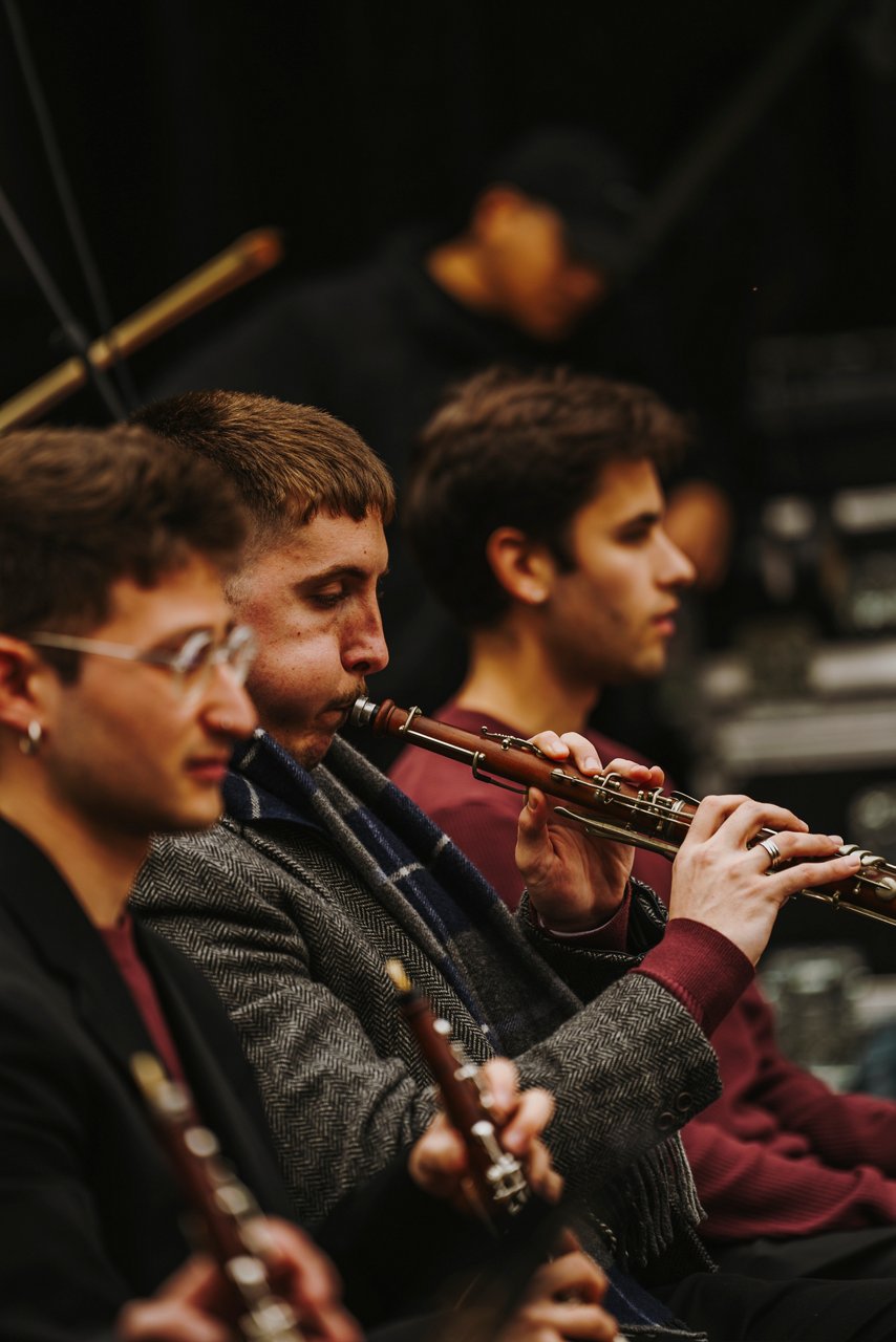 Close-up of three young musicians playing gralles, woodwind instruments used in sardanas and castells. They have a focused expression and look ahead. In the background, another musician appears out of focus on a dark stage.