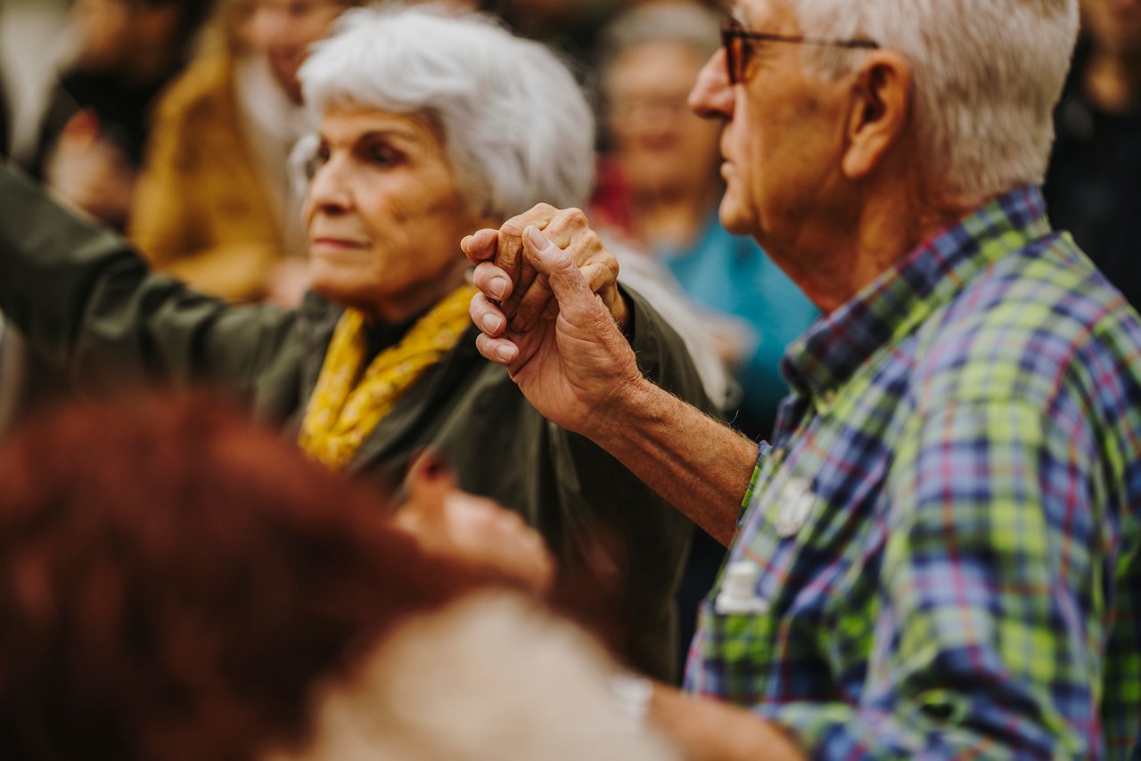 Close-up of an elderly couple dancing sardanas. The man is wearing a checkered shirt and the woman a yellow scarf around her neck. In the background, other people can be seen taking part in the dance, creating a sense of tradition and community.
