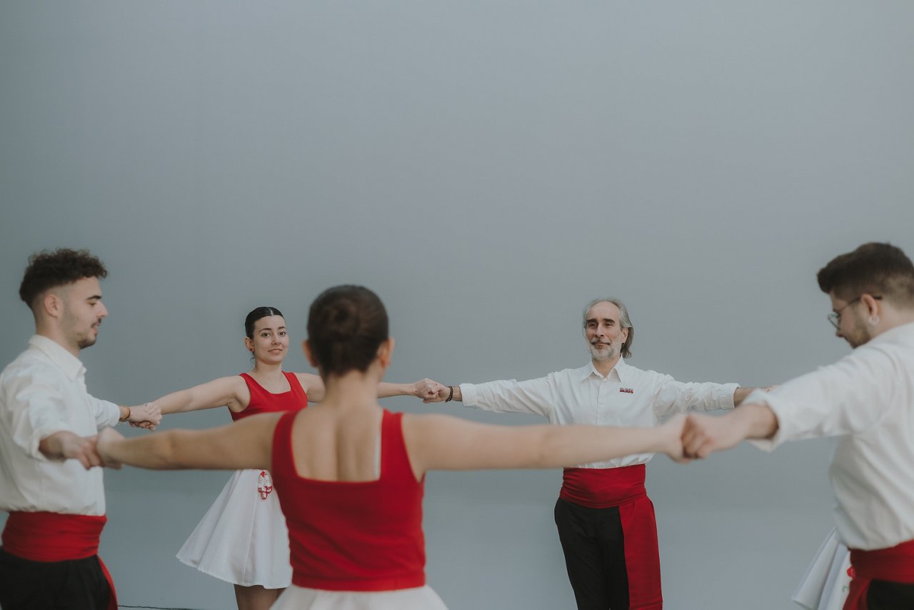 Group of people forming a circle and dancing sardanas in an indoor space with a neutral background. They wear traditional red and white attire and keep their arms extended and linked while performing the dance in coordination.