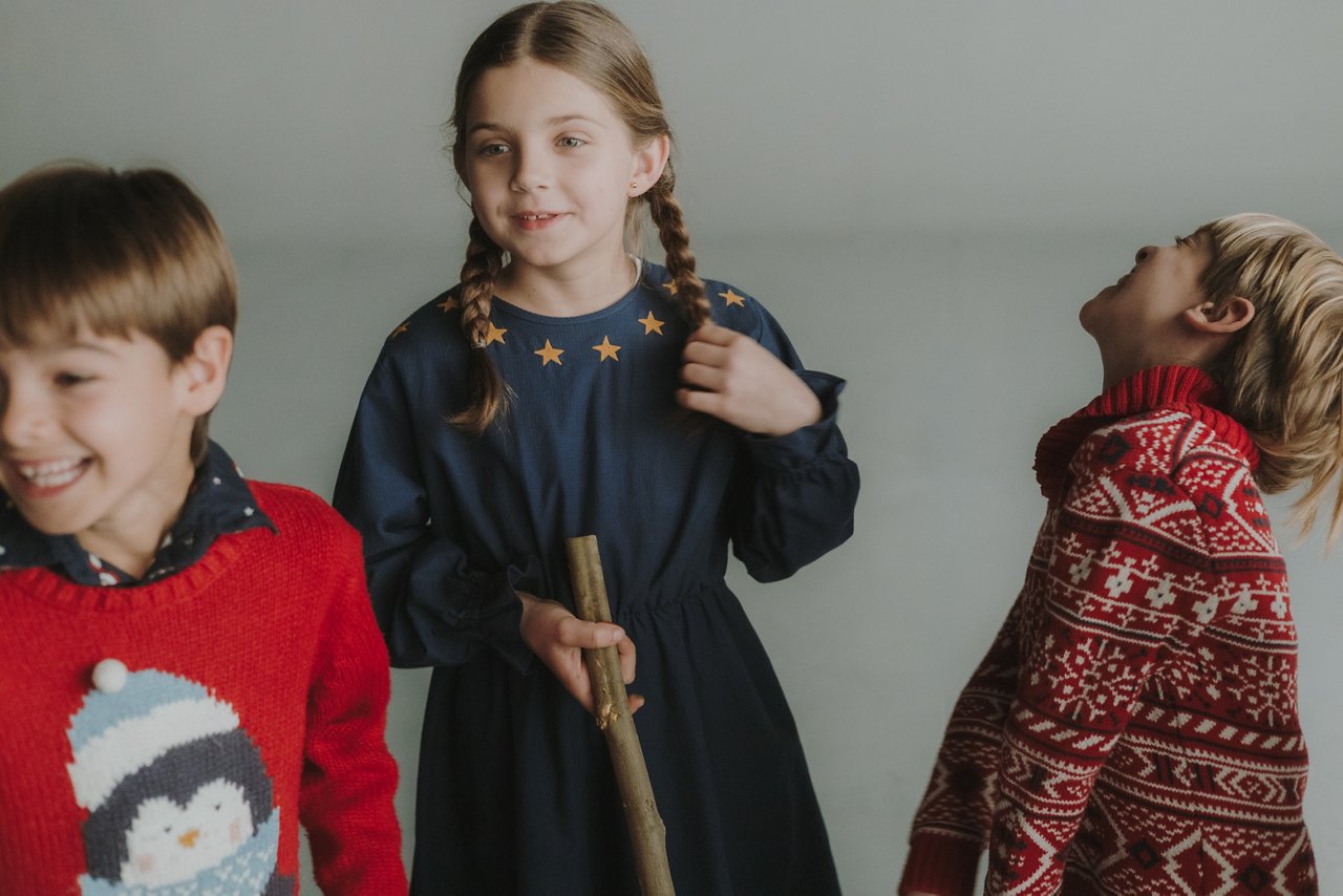 Three children wearing winter clothes and Christmas sweaters play and laugh, while a girl holds a stick in a home setting.