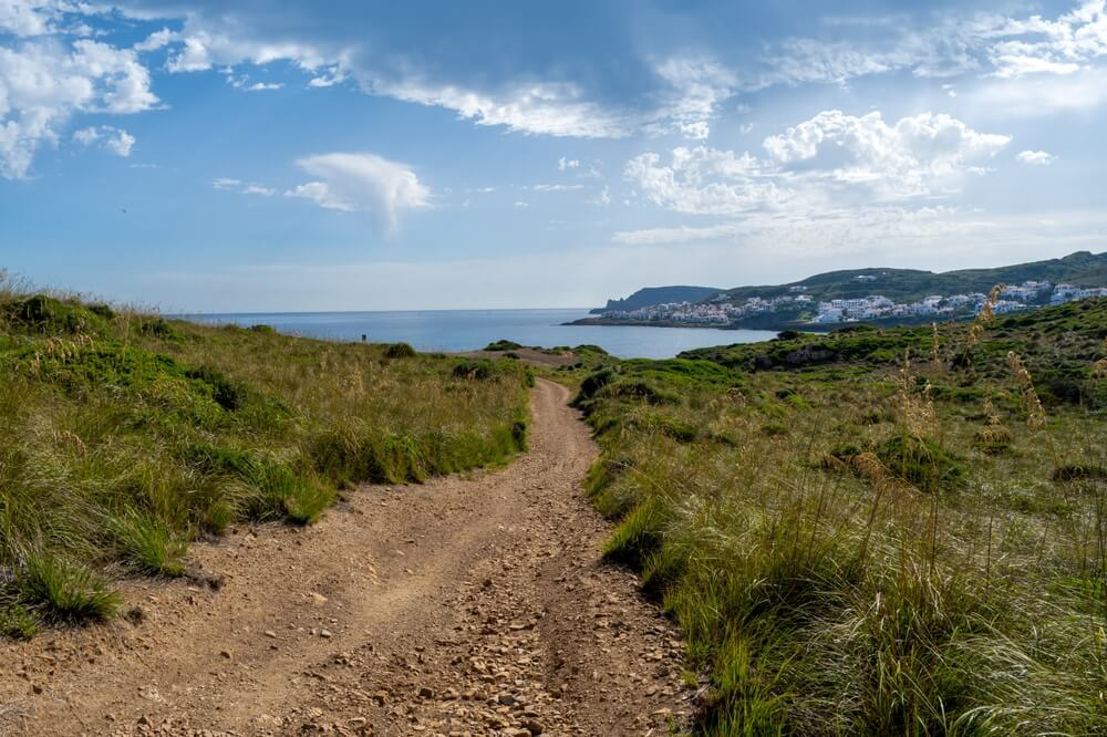 Menorca-Unternehmungen: Wanderweg in der Nähe der Küste mit einem Dorf im Hintergrund.