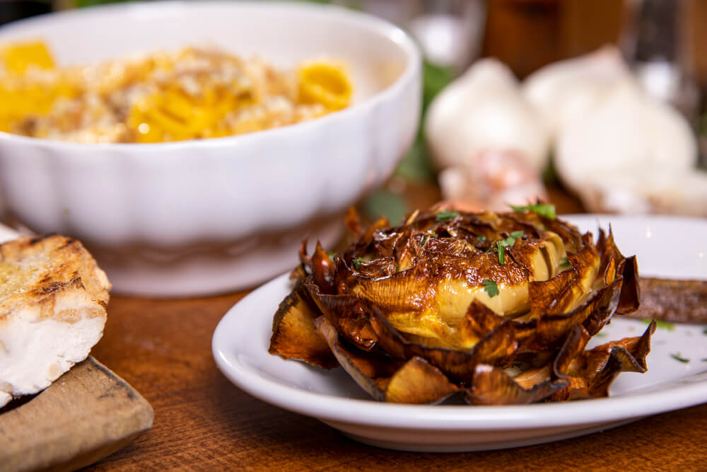 Carciofi alla Giudia: A rose shaped artichoke roasted on a white plate on a wooden table