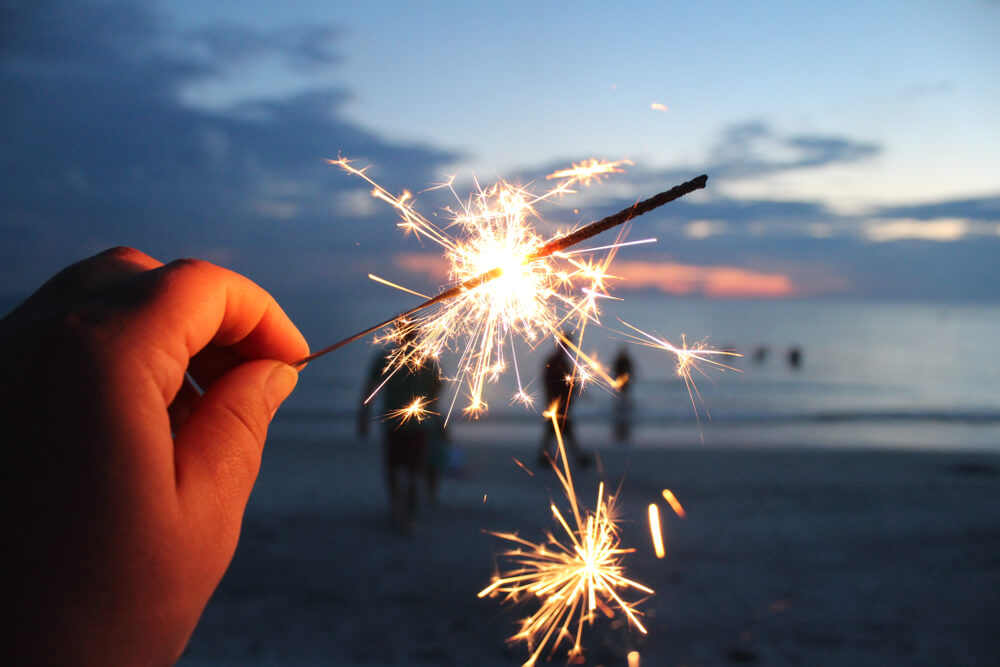 Hand mit Wunderkerze am Strand mit Menschen im Hintergrund.