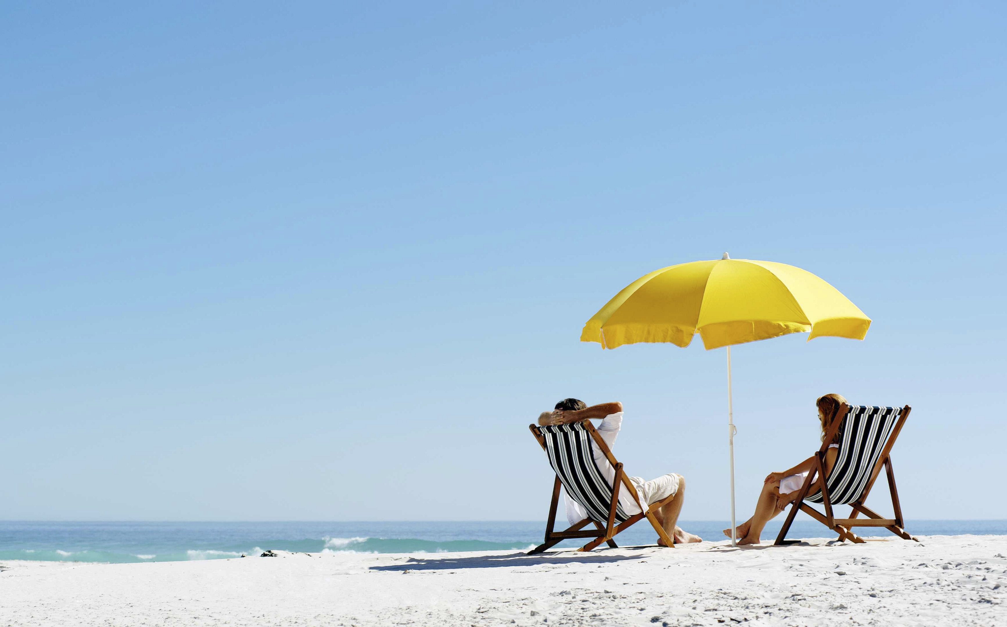 Beach summer couple on island vacation holiday relax in the sun on their deck chairs under a yellow umbrella. Idyllic travel background.