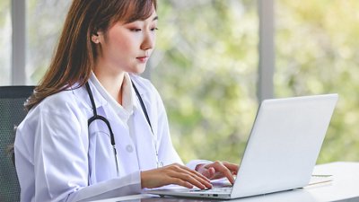 Educational Booklets Asian successful professional young female doctor in a white lab coat with a stethoscope, sitting and smiling while typing and working on a laptop.