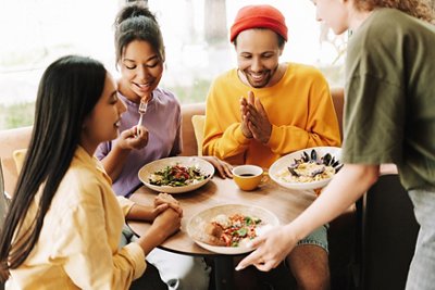 Customers enjoying a meal in a modern cafe, chatting happily. A diverse group of friends sharing salads and pasta, with natural light streaming in