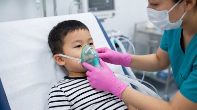 A child sits on a hospital bed while a healthcare worker places an oxygen mask on the child’s face. Medical equipment can be seen in the background. The worker is focused on providing care.