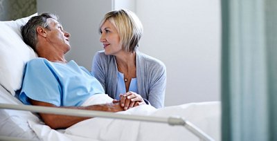 Elderly man in blue hospital gown in a hospital bed holds hands with and looks at woman visitor 