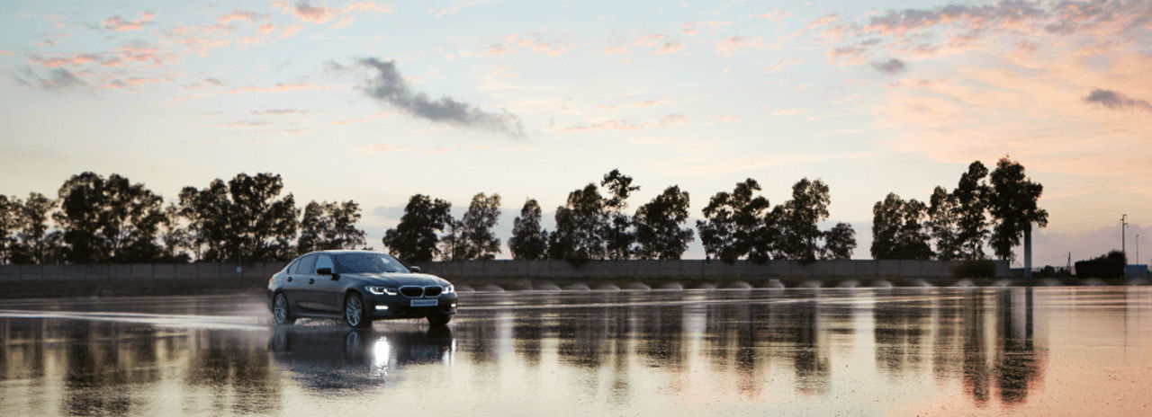 Un automóvil salpicando agua, reflejado sobre una pista mojada flanqueada de árboles y con una puesta de sol de fondo.
