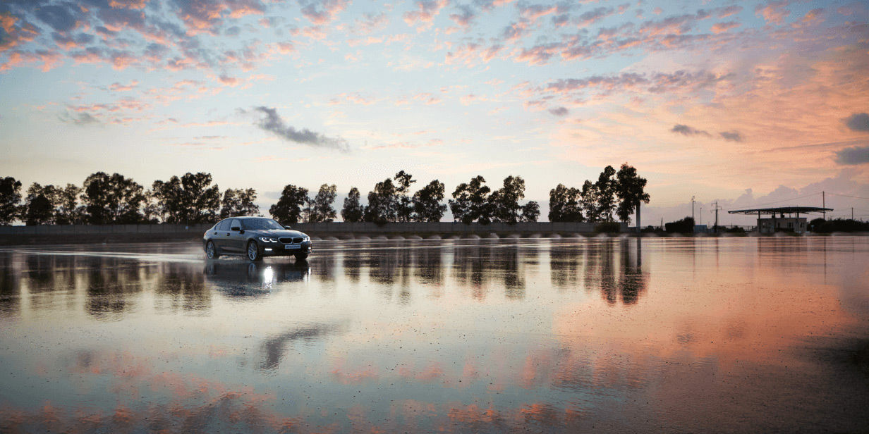 Un vehículo desplazándose en la distancia sobre una pista mojada flanqueada de árboles y con una puesta de sol de fondo.