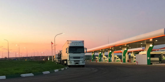 Deze afbeelding toont een vrachtwagen bij een tankstation langs de snelweg, met een zonsondergang op de achtergrond.