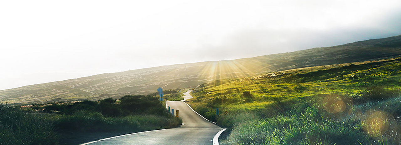 Eine lange Straße schlängelt sich um einen Berg mit einer malerischen grünen Weide und einem Sonnenuntergang.
