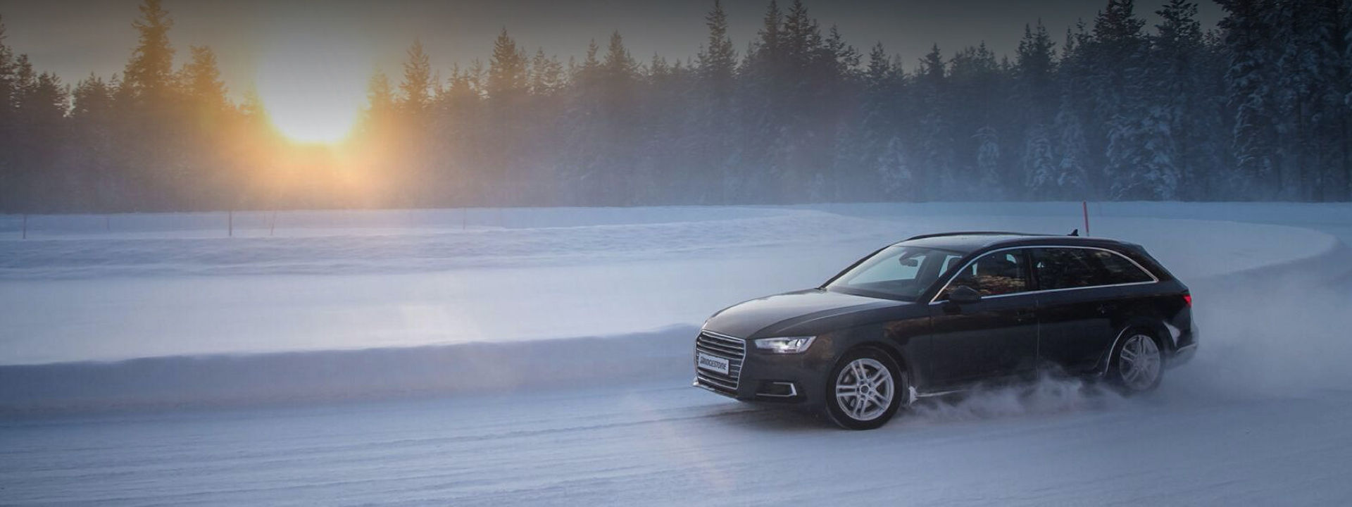 Een zwarte Audi uitgerust met Blizzak LM005-winterbanden draait de hoek om rond een sneeuwbank op een besneeuwde weg in het midden van een bos. 