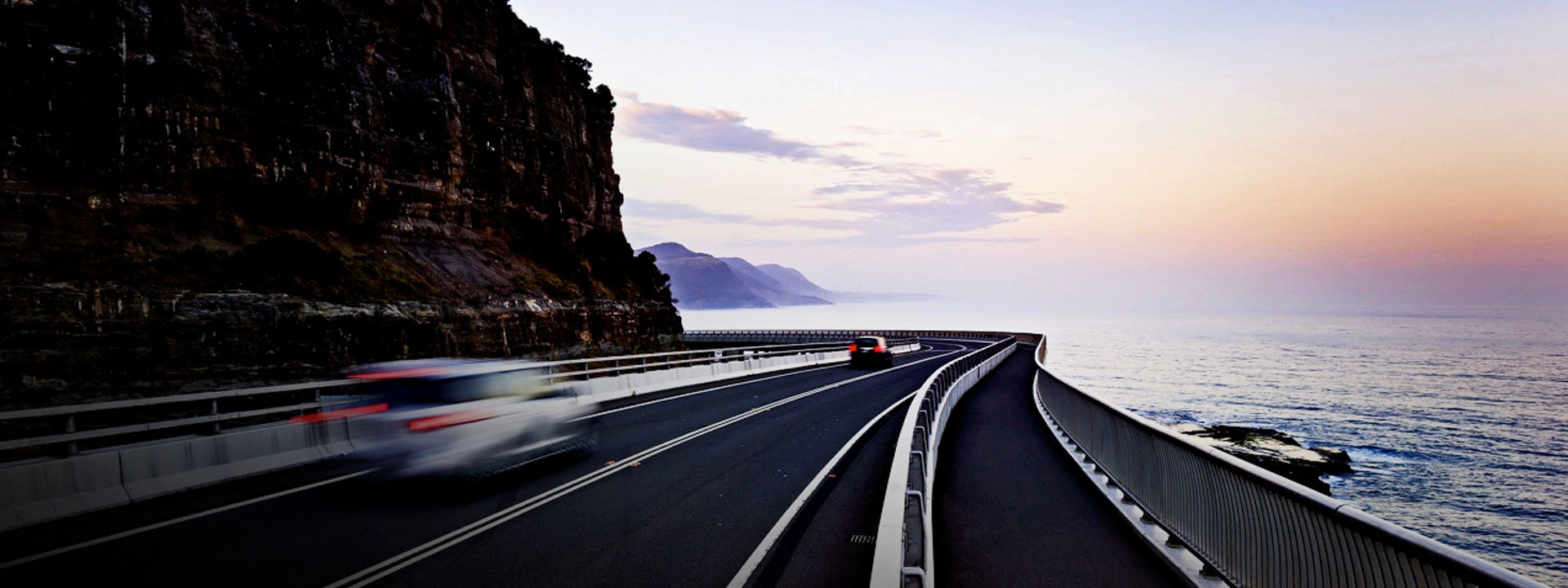 Cars equipped with Bridgestone tyres driving on a coastal highway with a sunset in the background.  