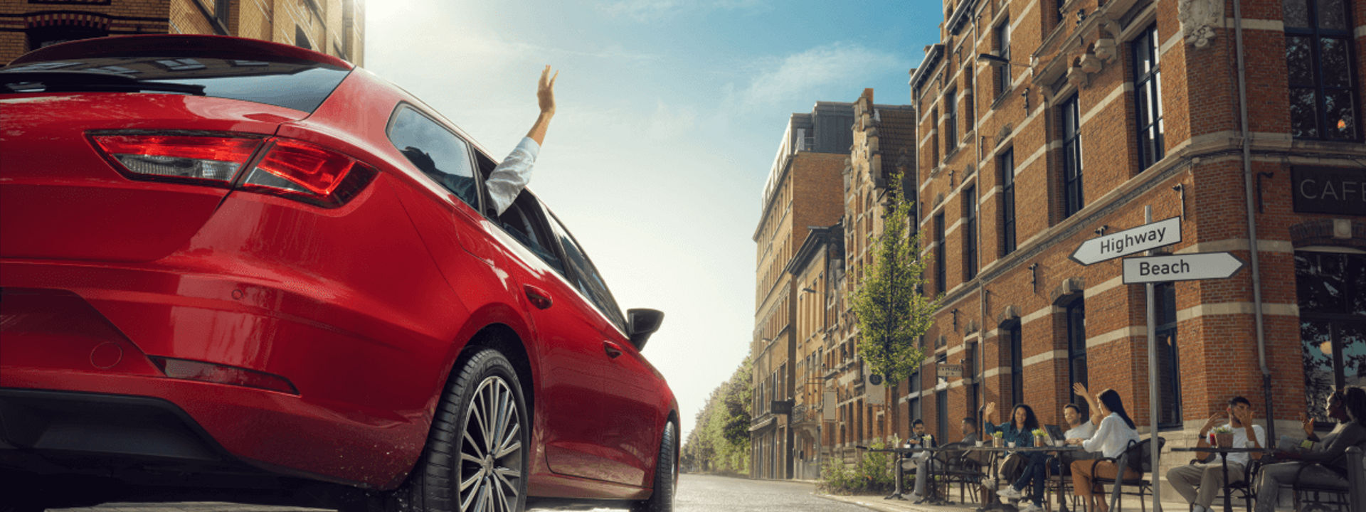 Ein rotes Auto mit Firestone-Roadhawk-2-Reifen fährt auf einer nassen Straße durch eine Altstadt und passiert dabei ein Straßencafé.