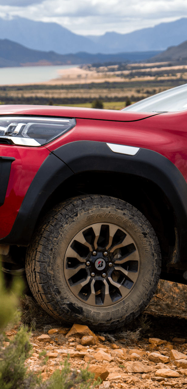 Raudono „Toyota Hilux“ su „Dueler All-Terrain A/T002“ padangomis, važiuojančio į kalną ant žvyro ir akmenų, vaizdas iš šono.