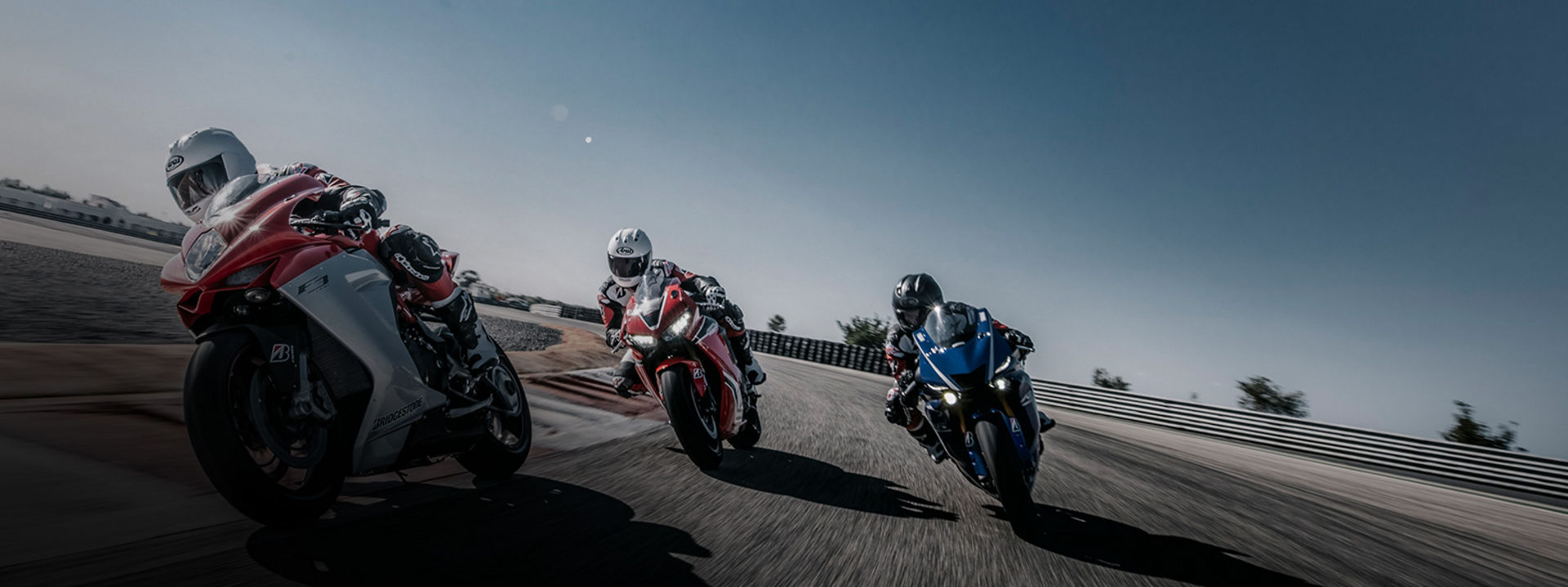 Na tej sliki so prikazani trije dirkači motornih koles v zavoju dirkališča pri visoki hitrosti s pnevmatikami Bridgestone. 