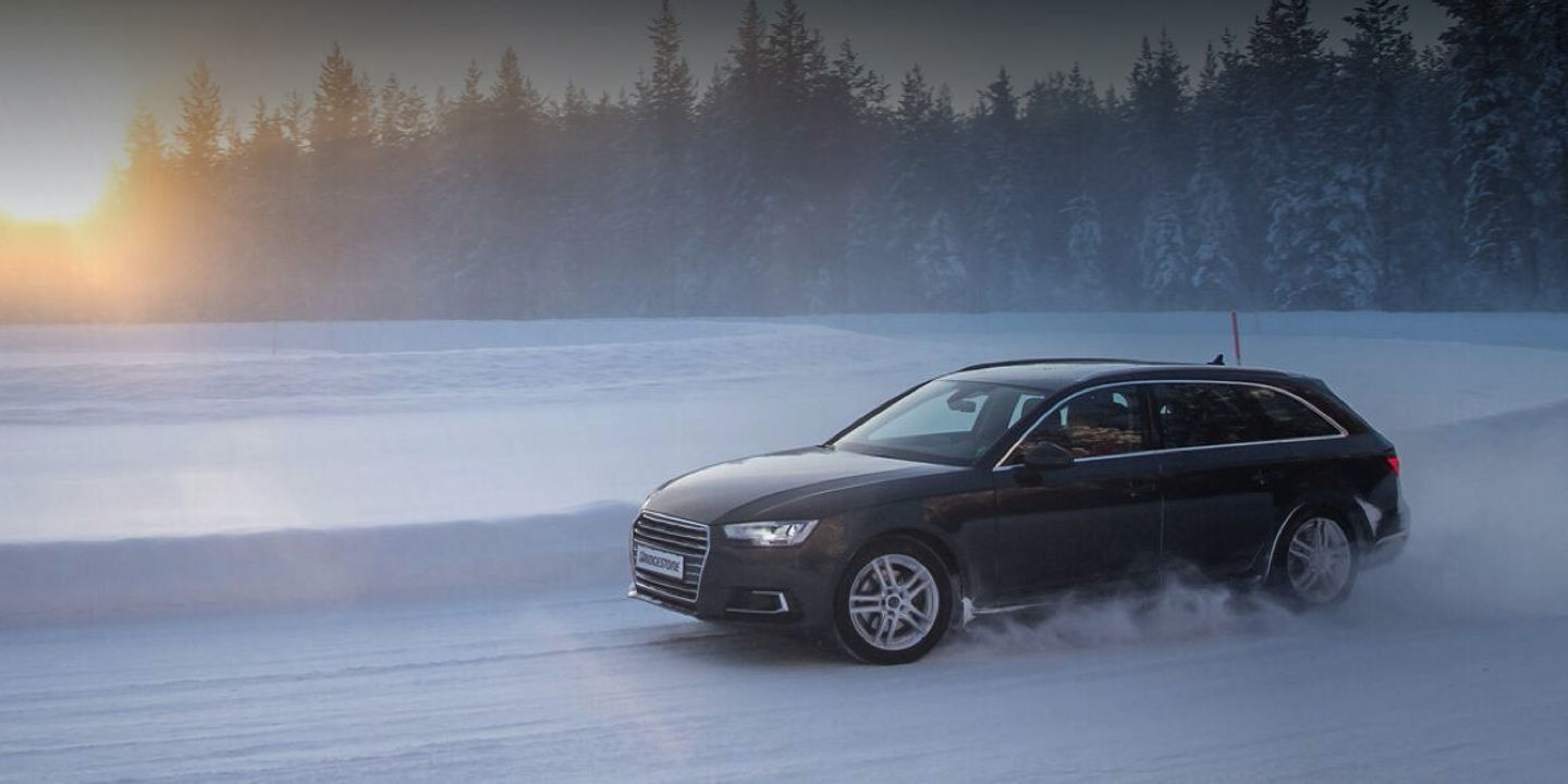 A black Audi equipped with Blizzak LM005 winter tyres is turning a corner around a snowbank on a snowy road in the middle of a forest. 