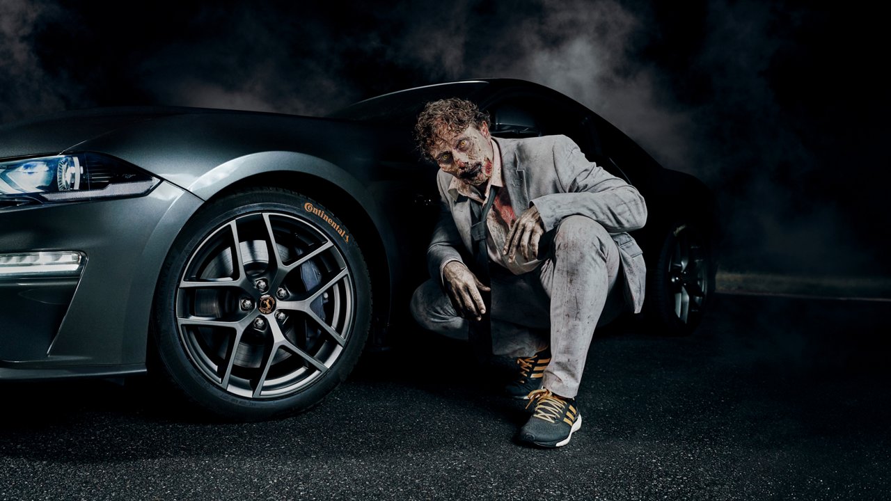 A zombie, wearing a grey suit, is crouching next to the Continental front tire.