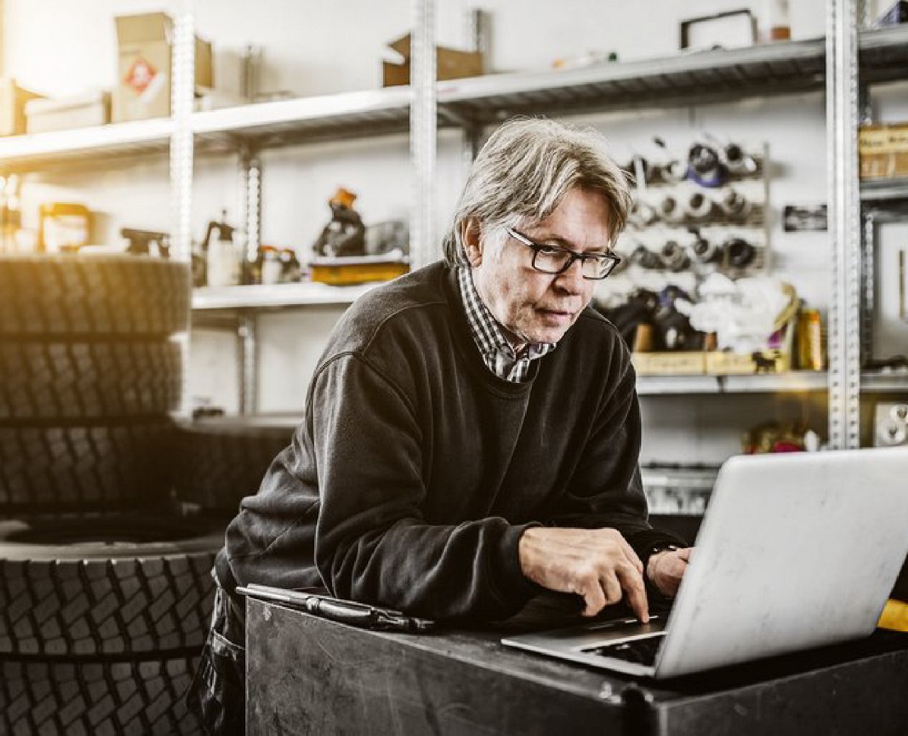 Older man working on a laptop in a workshop with tools, tires, and equipment in the background.