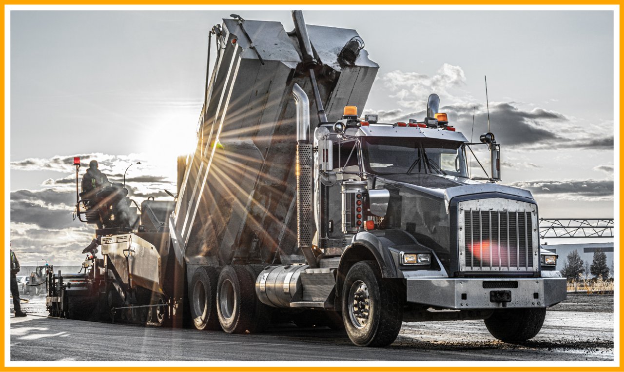 Dump truck unloading asphalt into a paving machine on a sunny road construction site.