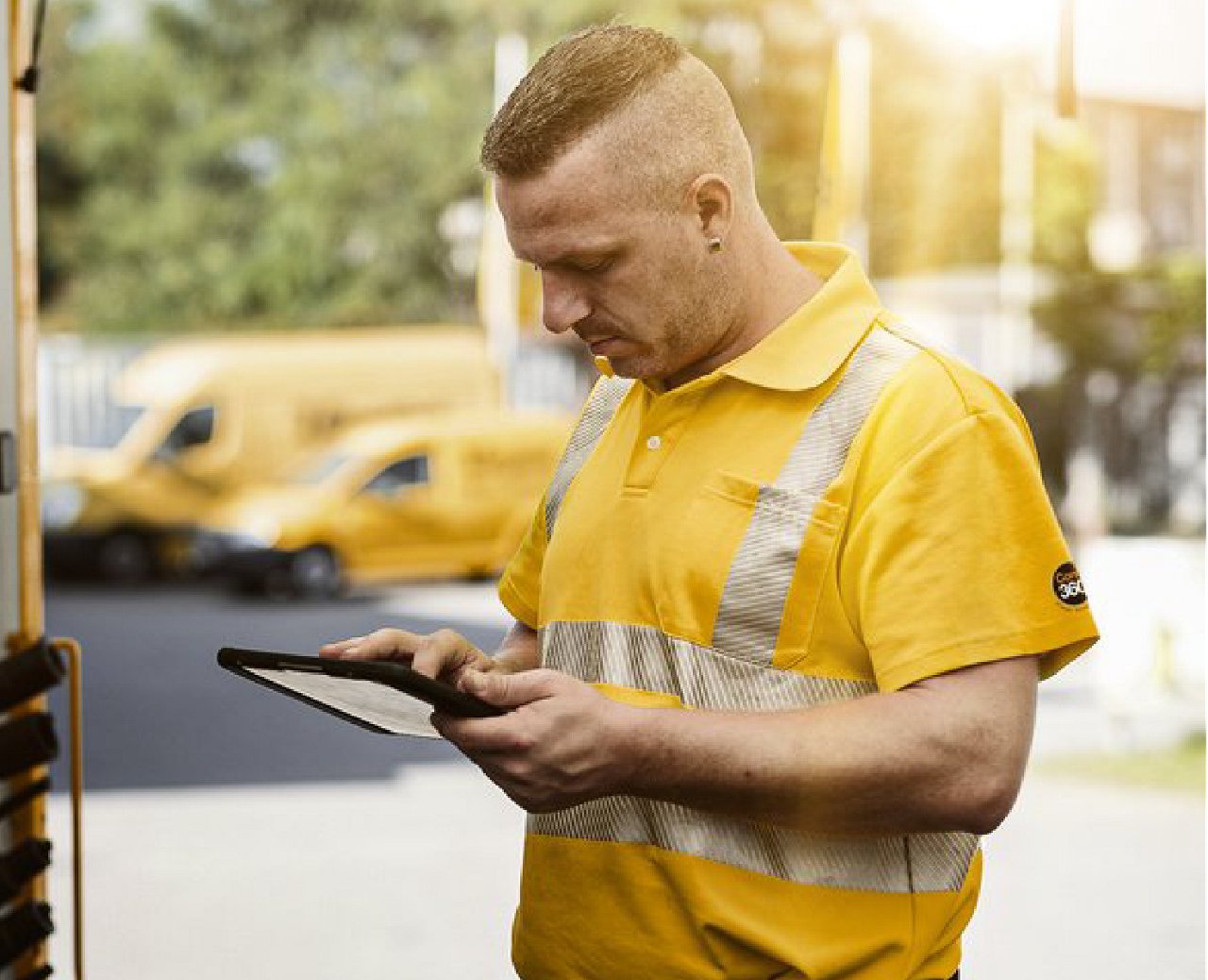 Worker in safety-striped shirt using a tablet near parked yellow delivery vans.