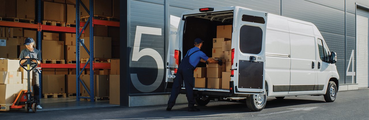 Workers load and move boxes at a warehouse with a delivery van parked outside.