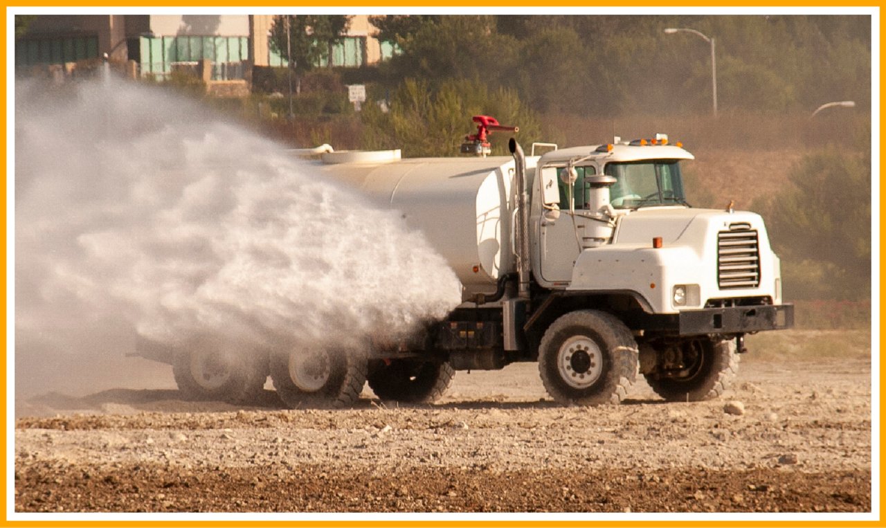 White water truck spraying mist onto dry ground, with trees and buildings in the background