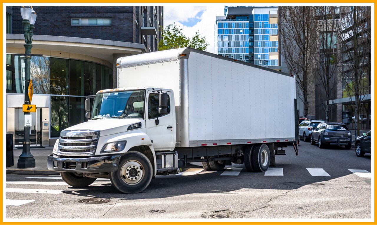 White box truck driving through a city intersection with buildings and trees in the background.