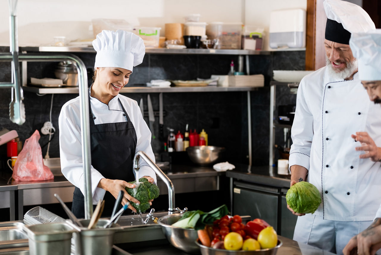 Smiling chef washing broccoli near interracial colleagues talking in restaurant kitchen; Shutterstock ID 2079121255; purchase_order: sito; job: ; client: ; other: 