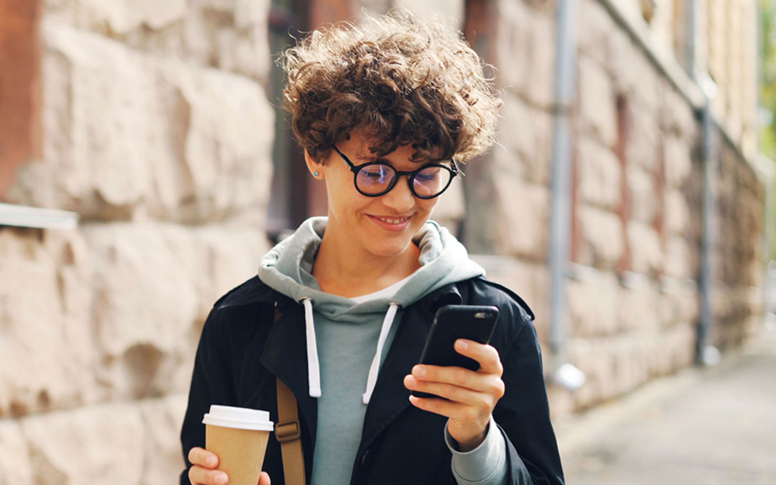 Eine Person läuft mit Coffee-to-go eine Straße entlang und schaut aufs Smartphone