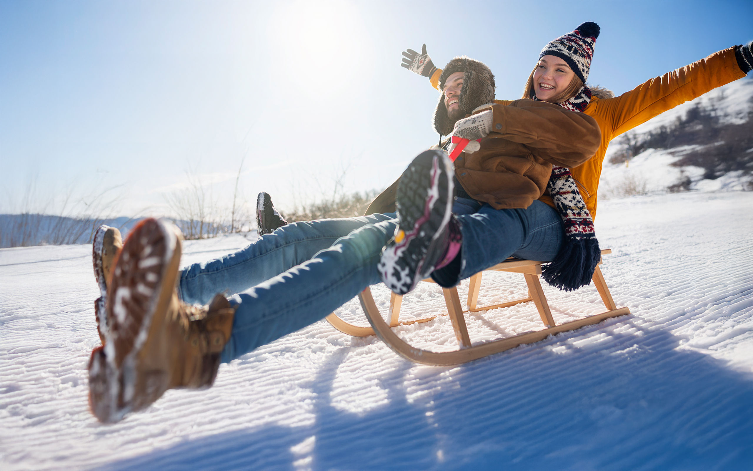 Ein junges Paar fährt vergnügt auf einem Schlitten bei Sonnenschein einen verschneiten Abhang hinab.