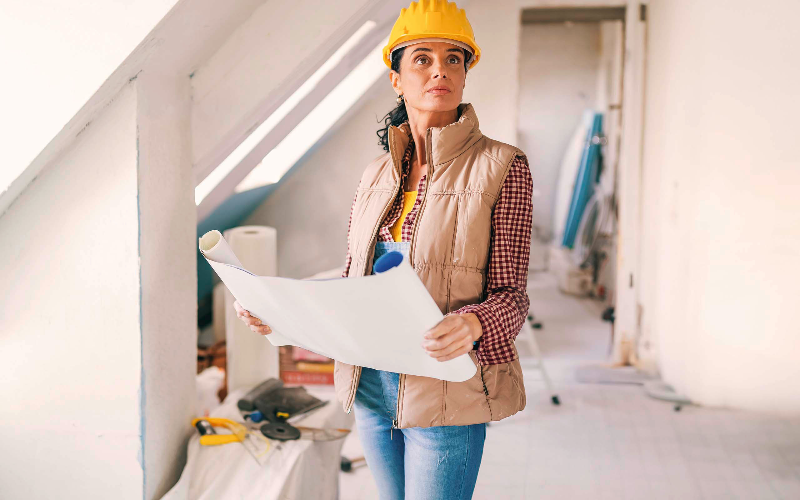 Eine Frau mit Bauhelm und Bauplan inspiziert ein Dachgeschoss, welches gerade ausgebaut wird