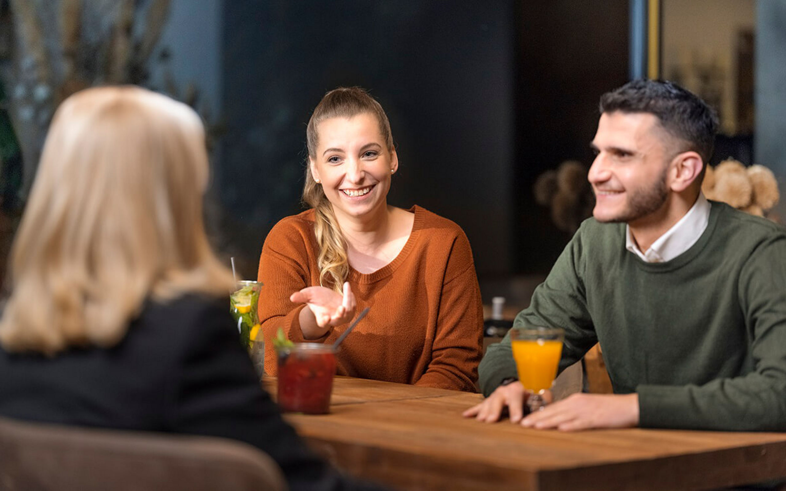 Drei Personen sitzen in einer Bar und unterhalten sich. Auf dem Tisch stehen Getränke..