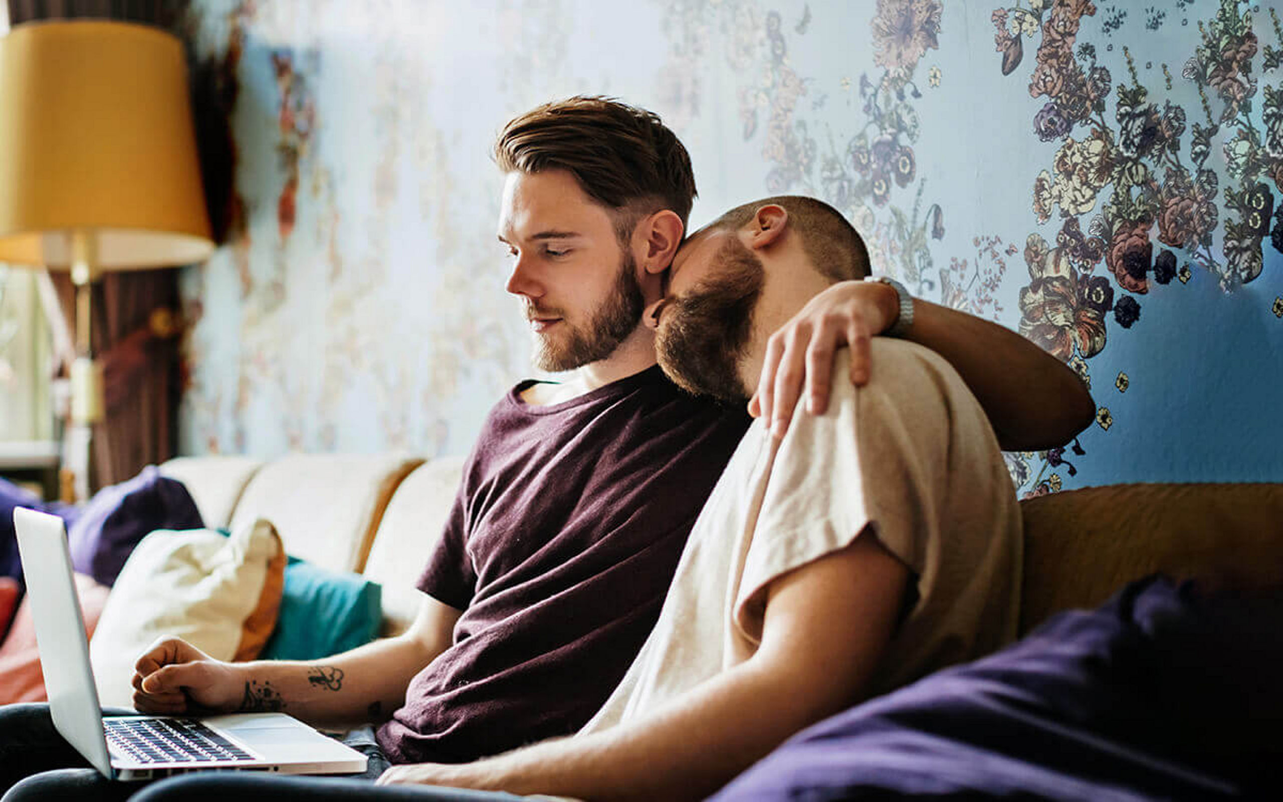 Zwei junge Männer sitzen Arm in Arm auf einer Couch und sehen auf einen Laptop