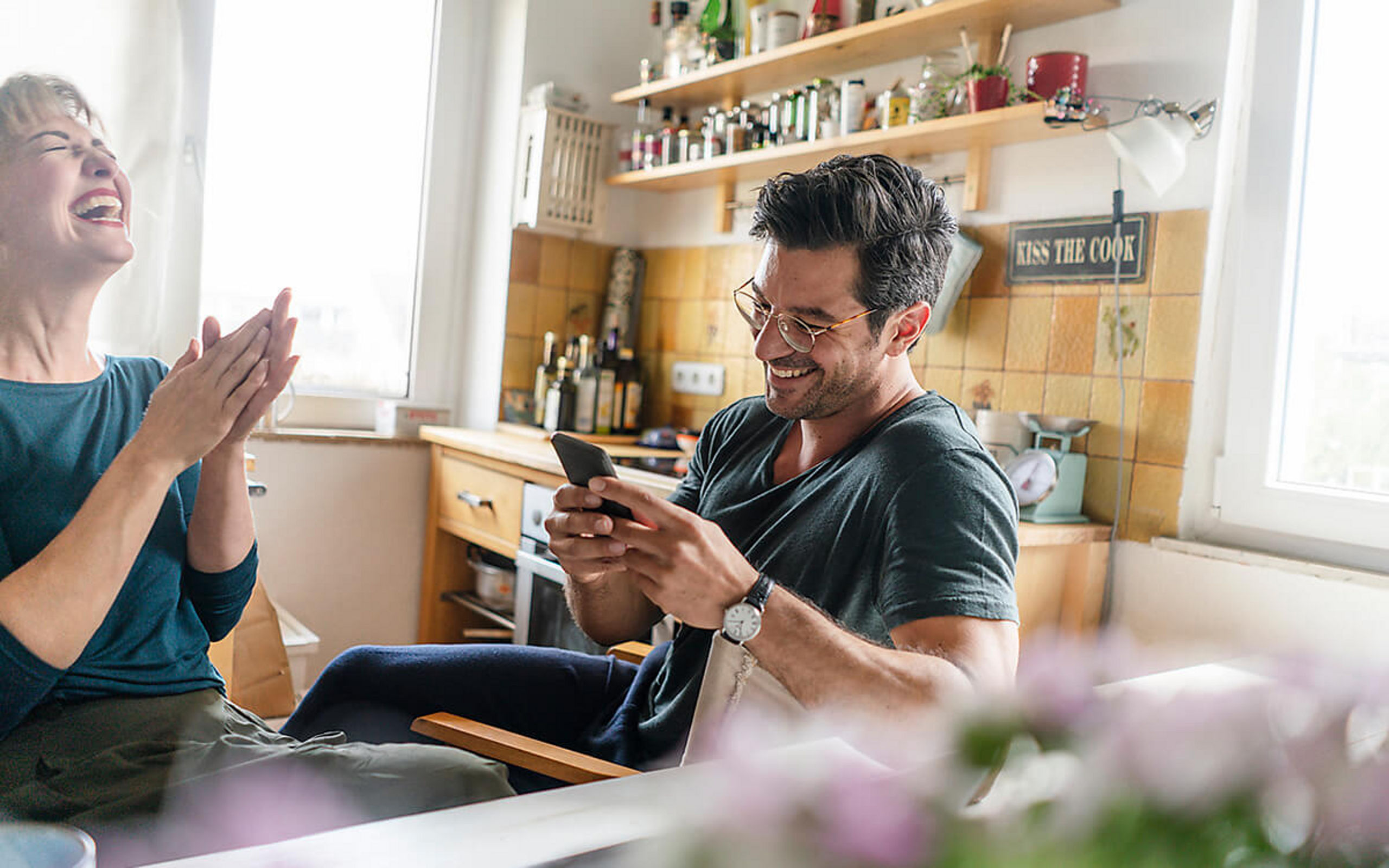 Ein junges Paar sitzt in der Küche und freut sich, während er auf sein Handy sieht