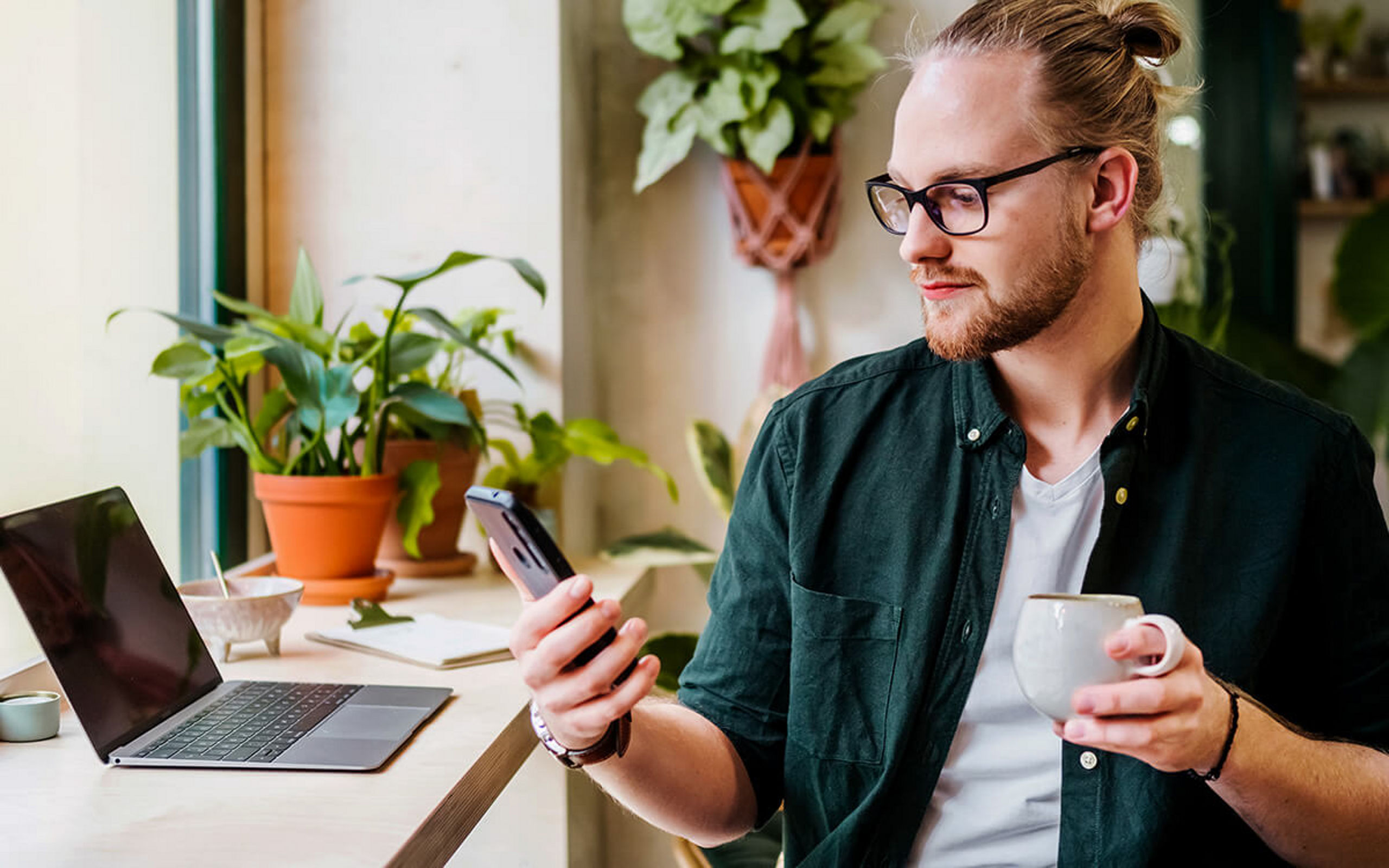 Eine Mann mit Kaffeetasse in der Hand sitzt vor einem Laptop und sieht auf sein Handy