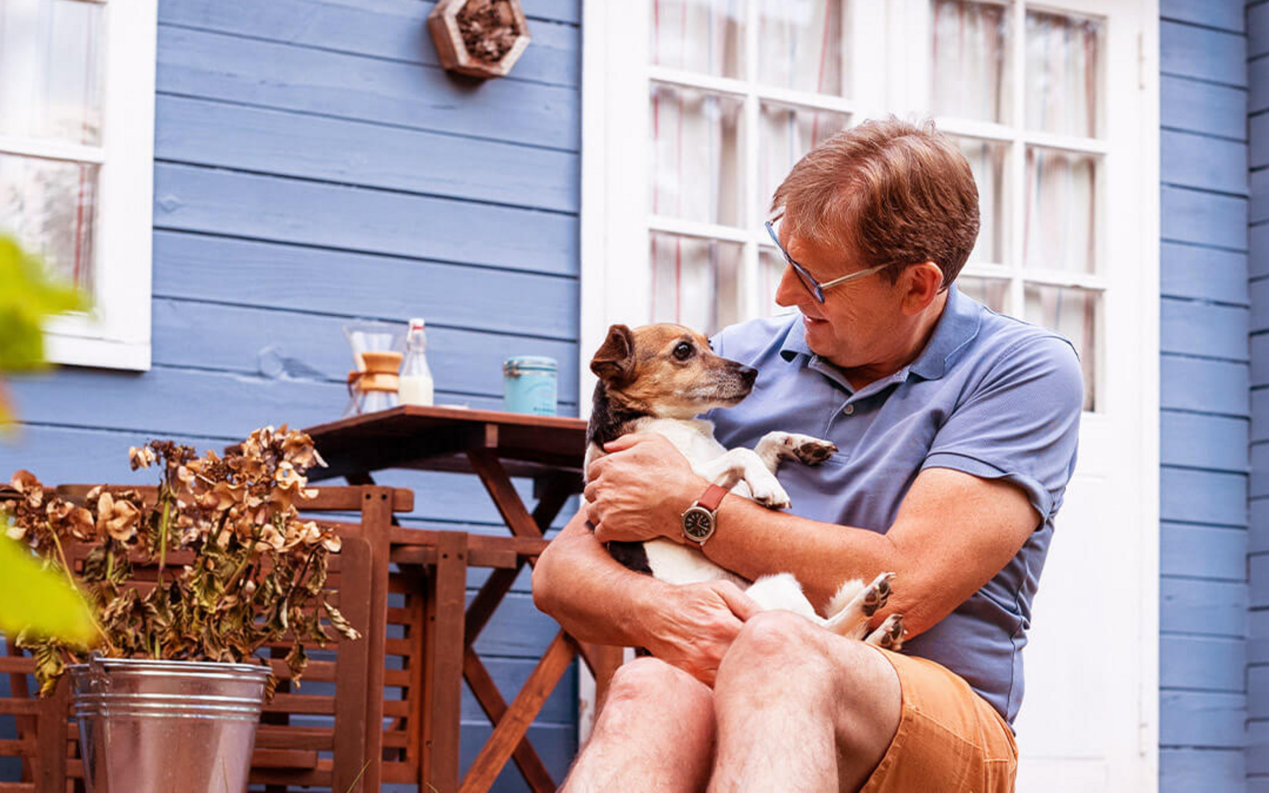 Eine Mann sitzt mit seinem Hund im Arm vor einem Gartenhaus